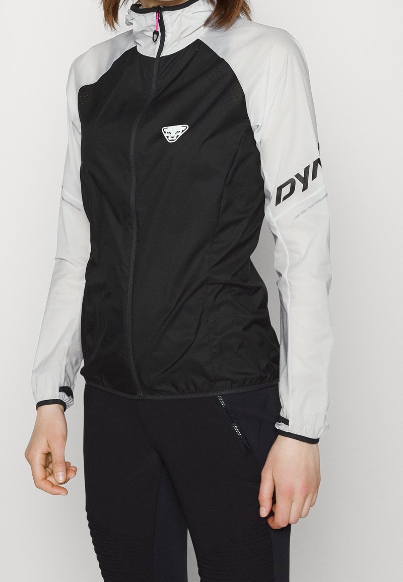 Veste légère noir et blanc avec une fermeture éclair, un col montant, des aérations discrètes et un logo sur la poitrine et la manche.