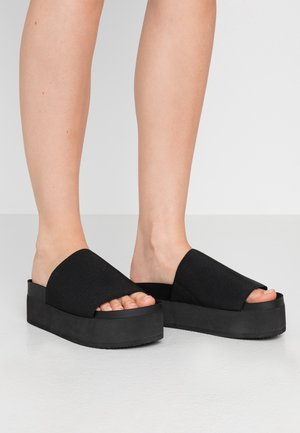 Beine mit schwarzen Plateau-Slide-Sandalen mit dicken Sohlen und breiten Stoffriemen über den Zehen auf einem hellgrauen Boden.