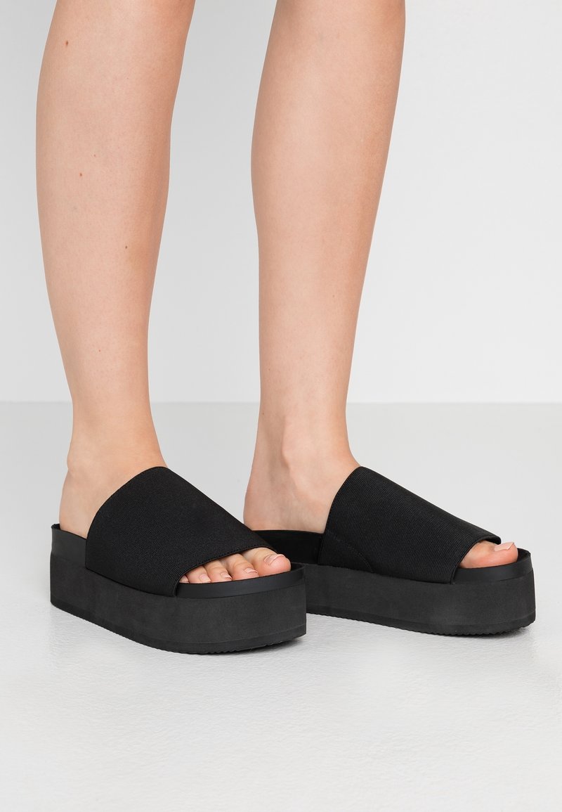 Jambes portant des sandales plates-formes noires avec semelles épaisses et larges brides en tissu sur les orteils sur un sol gris clair.