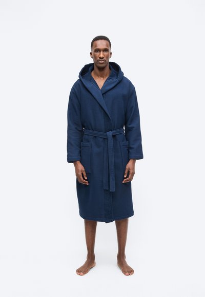 Άνδρας που στέκεται ξυπόλητος, φορώντας ένα σκουρομπλε μπουρνούζι με κουκούλα μέχρι το γόνατο, με ζώνη και μπροστινές τσέπες, μπροστά σε απλό φόντο.