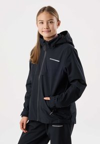 Mlada oseba, ki nosi črno jakno s kapuco in ujemajoče se hlače, stoji z eno roko v žepu, nevtralna svetla podlaga.