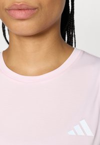 Αθλητική ροζ μπλούζα με υφή υφάσματος, που φέρει λευκό λογότυπο Adidas στο κάτω δεξί μέρος. Στρογγυλή λαιμόκοψη, κοντά μανίκια.