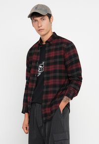 Joven con una gorra gris, camisa de cuadros burdeos y negra sobre una camiseta gráfica negra, pantalones cargo gris oscuro y un tatuaje en el antebrazo izquierdo.