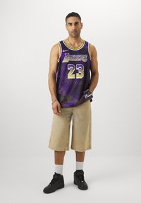 Maglia viola dei Los Angeles Lakers con dettagli gialli e numero 23, indossata sopra pantaloni beige, abbinata a sneaker nere alte e calze bianche.