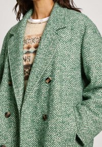Double-breasted green herringbone coat with a soft texture; features large brown buttons and a notched collar, layered over a patterned sweater.