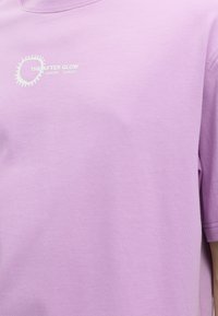 Λιλά κοντομάνικη μπλούζα με λευκή επιγραφή "THE AFTER GLOW SAMSOE SAMSOE" και γραφικό ήλιου στην πάνω αριστερή πλευρά του στήθους.