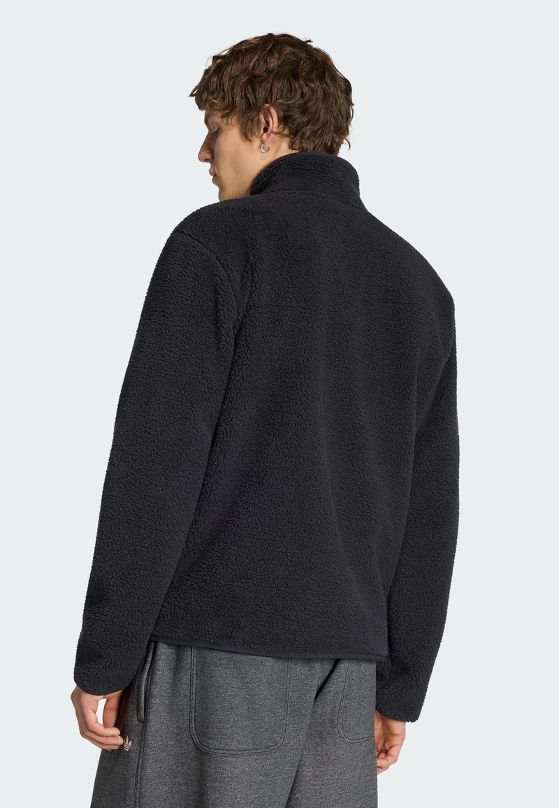 Homme portant une veste polaire noire à col montant, vu de dos, associé à un pantalon décontracté gris foncé.