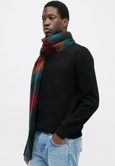 Uomo che indossa un maglione nero, jeans blu e una sciarpa di lana a righe colorate avvolta attorno al collo, in piedi con una mano in tasca.