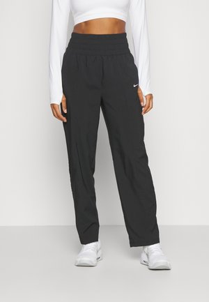 Frau trägt schwarze Nike Sporthose mit hohem Bund, weißes kurz geschnittenes Langarmshirt und weiße Sneaker, steht vor hellem Hintergrund.