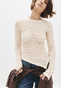 Vrouw in een crèmekleurige kant lange mouwen top en blauwe spijkerbroek, met een donkerbruine blouse die om haar taille is geknoopt, voor een effen witte achtergrond.