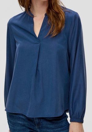 Mujer con una blusa azul marino de manga larga con escote en V y detalle plisado, combinada con vaqueros de mezclilla azules.