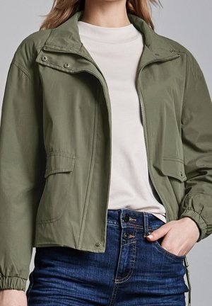 Femme portant une veste zippée vert olive sur une chemise blanche et un jean bleu taille haute, main dans la poche.