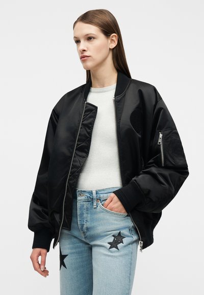 Νέα γυναίκα που φοράει μαύρο μπουφάν τύπου bomber, ανοιχτό γκρι ριμπ τοπ και ανοιχτό μπλε τζιν με μαύρα αστέρι επιθέματα, με το χέρι στην τσέπη, ουδέτερο φόντο.