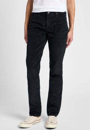 Osoba nosící černé manšestrové kalhoty, bílé tenisky a bílé tričko stojí před jednobarevným světlým pozadím.