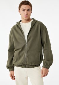 Chaqueta verde oliva con cremallera frontal y capucha, puños y cinturilla elásticos. Presenta una textura suave y bolsillos laterales para mayor utilidad.