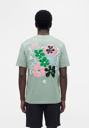 Moški nosi svetlo zeleno majico s pisanim rožnatim, zelenim in črnim cvetličnim vzorcem na hrbtu in črne kratke hlače, stoji obrnjeno stran.