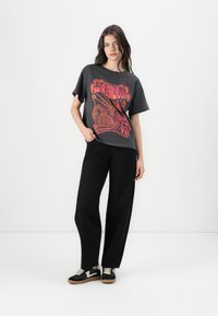 Grafisches schwarzes T-Shirt mit auffällig rotem Design, entspannte Passform, kurze Ärmel, kombiniert mit schwarzen weiten Hosen und schwarzen Turnschuhen mit weißen Akzenten.