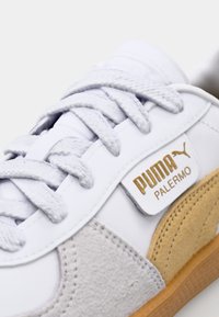 Bílé tenisky s šedými semišovými detaily, texturovanými tkaničkami a zlatou logovou nášivkou. Mají gumovou podešev a hladký kožený svršek.