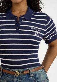 Sieviete valkā tumši zilu un baltu svītrainu Ralph Lauren polo kreklu, apvienotu ar zilām džinsu biksēm un brūnu jostu ar zelta sprādzi.