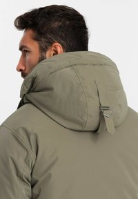 Khaki-gefütterte Jacke mit großer Kapuze, die mit einem Riemen und Metallschnalle verziert ist. Glatte Textur, strukturierte Silhouette.