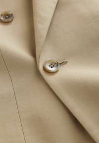 Dettaglio di una giacca in tessuto beige caratterizzata da una texture liscia, con un grande bottone rotondo e una leggera lucentezza. La cucitura è visibile lungo il bordo.