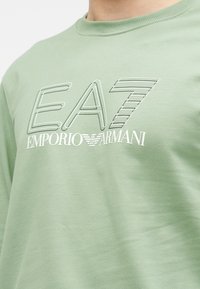 Camisa de algodón verde claro con un logo destacado de EA7 y el texto "Emporio Armani" en blanco debajo. Cuello redondo y textura suave.