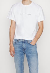 Hombre con una camiseta blanca que tiene el texto "La vida está justo fuera de tu puerta", combinada con jeans de un azul claro, de pie con las manos en los bolsillos.