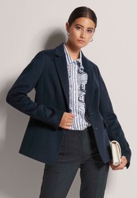 Marineblazer mit zwei Knöpfen und klassischem Revers, getragen über einer gestreiften, gerafften Bluse, kombiniert mit dunklen Hosen und gehalten mit einer leichten Clutch.