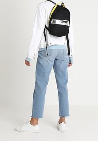 Mochila de malla negra con detalles en amarillo y logotipo plateado reflectante. Modelo vistiendo chaqueta blanca, suéter azul claro y jeans cortos.