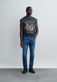 Mladý muž v sivom tielku s potlačou, modrých džínsoch, čiernom opasku a čiernych teniskách stojaci pred jednoduchou svetlosivou stenou.