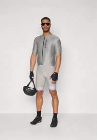 Tenue de cyclisme grise à manches courtes avec fermeture éclair, assortie à un short gris. Gants noirs, chaussures noires et casque noir, portés par un modèle masculin debout.