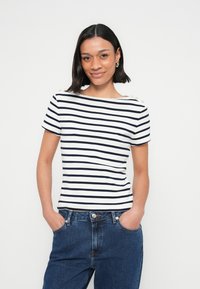 Ριγετό κοντομάνικο τοπ σε λευκό και ναυτικό μπλε, με λαιμόκοψη καραβοφάναρο και χρυσά κουμπιά διακοσμητικά στους ώμους. Συνδυάζεται με μπλε τζιν.