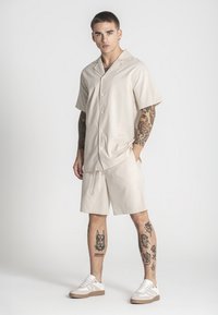 Camisa de manga corta color beige con botones, combinada con pantalones cortos a juego, ambos en un tejido texturizado. Zapatillas deportivas con parte superior blanca y detalles en beige.