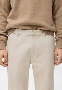 Pantalones beige hechos de tela suave, con cierre de botones, trabillas para cinturón y bolsillos laterales. Combinados con un suéter de punto marrón claro.