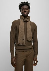 Pull en tricot marron avec une écharpe marron assortie comportant des bords à franges, placé sur un fond neutre.