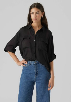 Camicia nera con bottoni, due tasche sul petto, maniche lunghe arrotolate. Indossata con jeans in denim blu a vita alta. Tessuto morbido, vestibilità comoda.
