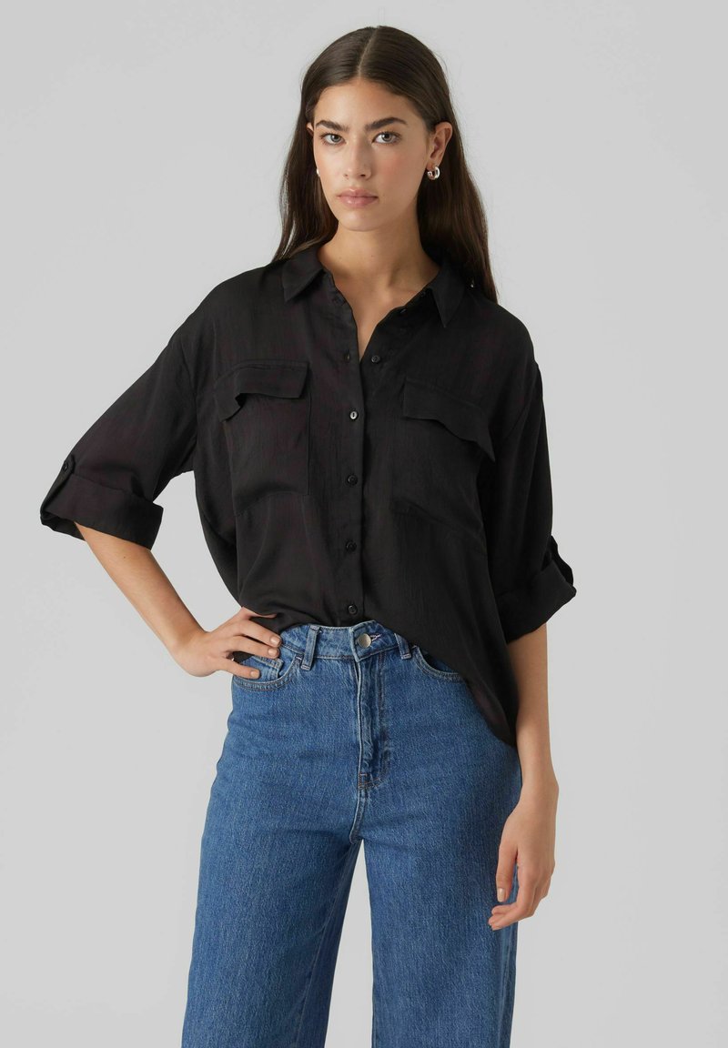 Camicia nera con bottoni, due tasche sul petto, maniche lunghe arrotolate. Indossata con jeans in denim blu a vita alta. Tessuto morbido, vestibilità comoda.