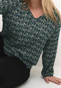 Haut à manches longues vert et crème avec un motif floral et un décolleté en V. Fabriqué en tissu doux, il présente une coupe décontractée et des poignets froncés.