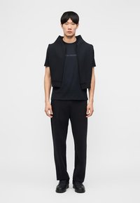 Νεαρός άνδρας φορώντας μαύρο μπλουζάκι, μαύρο παντελόνι, μαύρα αθλητικά παπούτσια και μαύρο φούτερ περασμένο στους ώμους, στέκεται μπροστά σε λευκό φόντο.