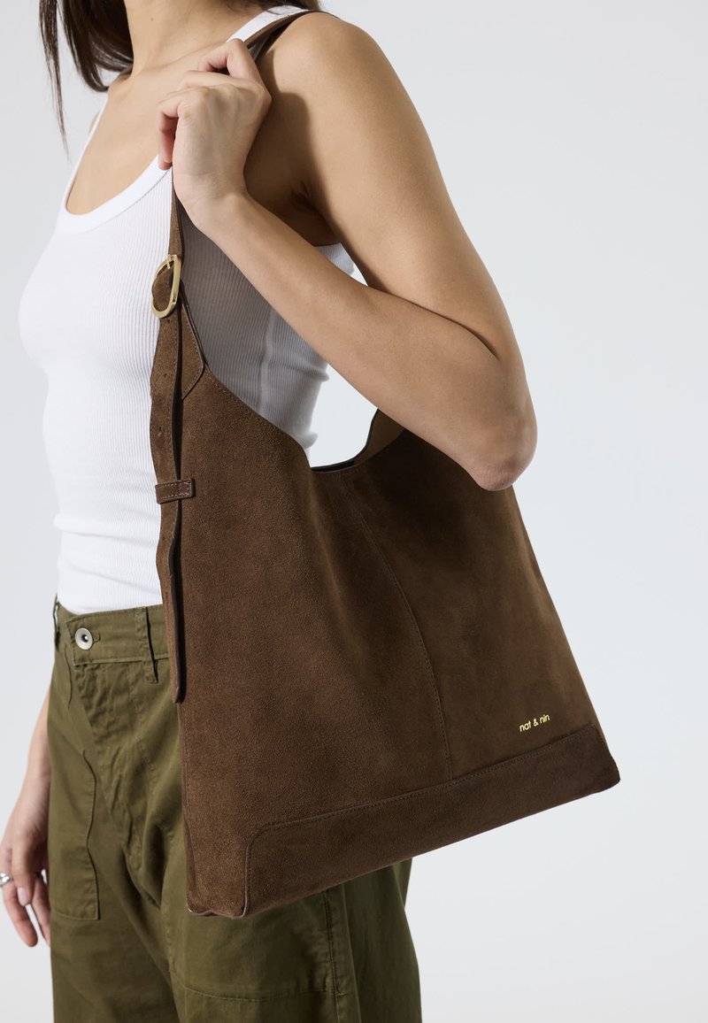 Femme portant un débardeur blanc et un pantalon olive, portant un grand sac à épaule en daim marron avec une boucle dorée et un petit logo.