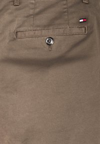 Bruine katoenen broek met een achterzak met een zilveren knoop en een klein logo-label in blauw, wit en rood aan de zijkant.