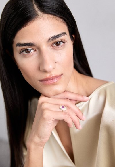 Goldener Ring mit lila Herzdetail, getragen an einer Hand, die auf dem Kinn ruht, ergänzt von einem satiniert beigen Oberteil und einer glatten Hautstruktur.
