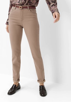 Personne portant un pantalon beige slim avec revers roulés, ceinture marron, chemise à motif cachemire et mocassins marron foncé, debout sur un fond blanc.