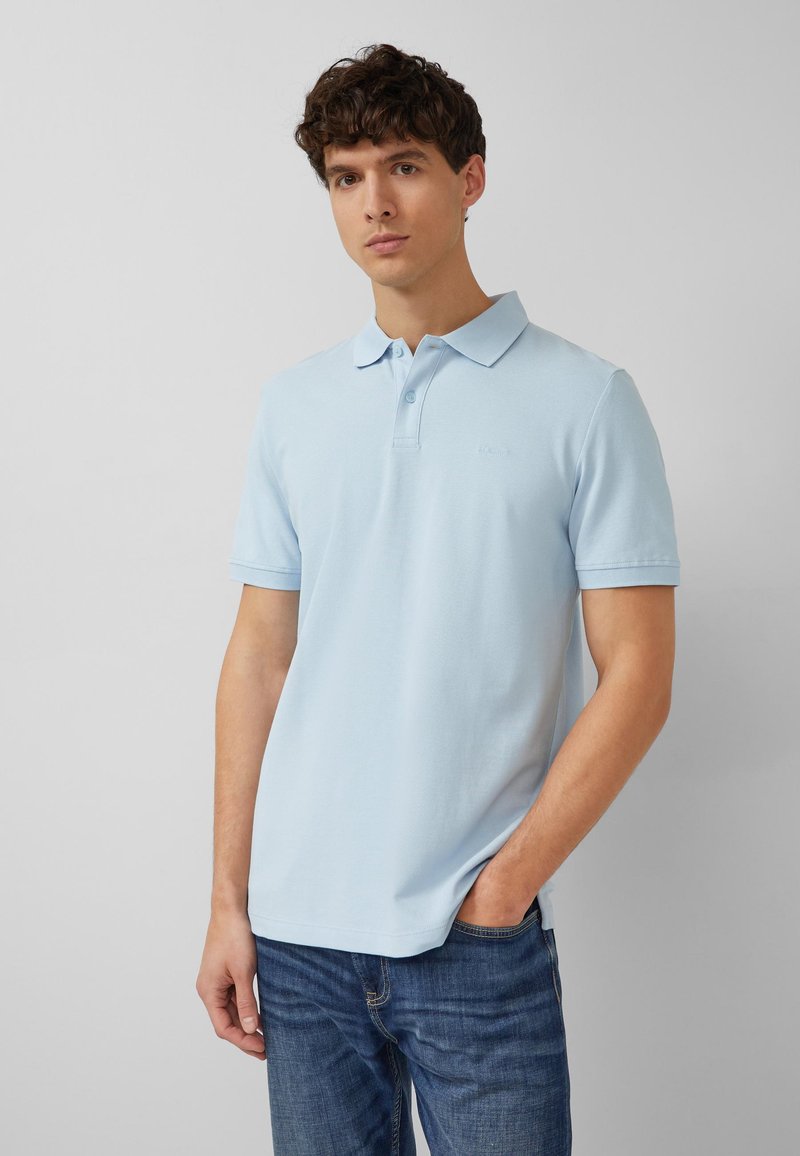 Giovane uomo con indosso una polo azzurro chiaro e jeans blu, in piedi davanti a uno sfondo grigio chiaro uniforme con una mano in tasca.