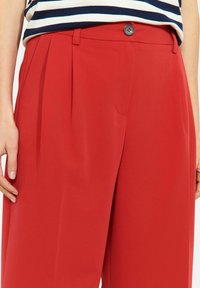 Punased püksid, millel on sile tekstuur, ees nööpidega kinnis, voldid ja lõikega istuvus. Osaliselt nähtav triibuline top.