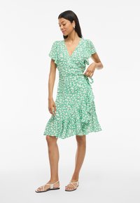 Grønn omslagskjole med v-hals, korte flommesermer og volangkant. Har et hvitt abstrakt mønster og en detalj med siden knytting.