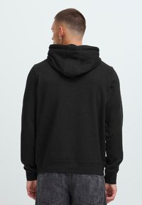 Uomo con capelli corti e orecchino, indossa un semplice hoodie nero e jeans neri sbiaditi, in piedi di spalle contro uno sfondo chiaro.