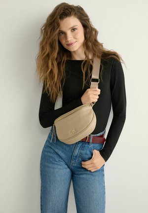 Kobieta o falowanych włosach, ubrana w czarną bluzkę z długim rękawem, niebieskie dżinsy i beżową torbę na ramię, trzymająca pasek torby, stojąca na tle jasnej, gładkiej ściany.