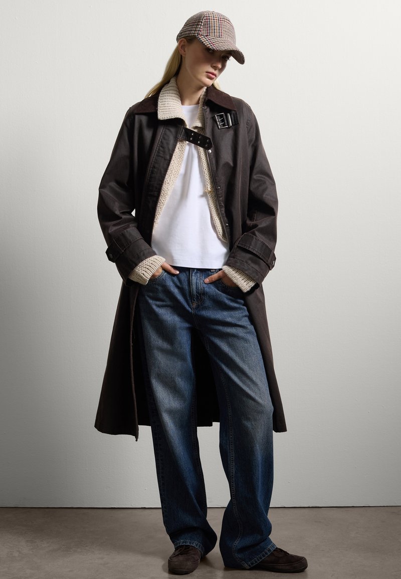 Manteau trench marron avec col en tricot beige, T-shirt blanc, jean large bleu et casquette à carreaux. Des chaussures marron complètent la tenue.