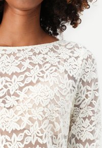 Haut en dentelle blanche avec des motifs floraux complexes, un col bateau et une texture semi-transparente.